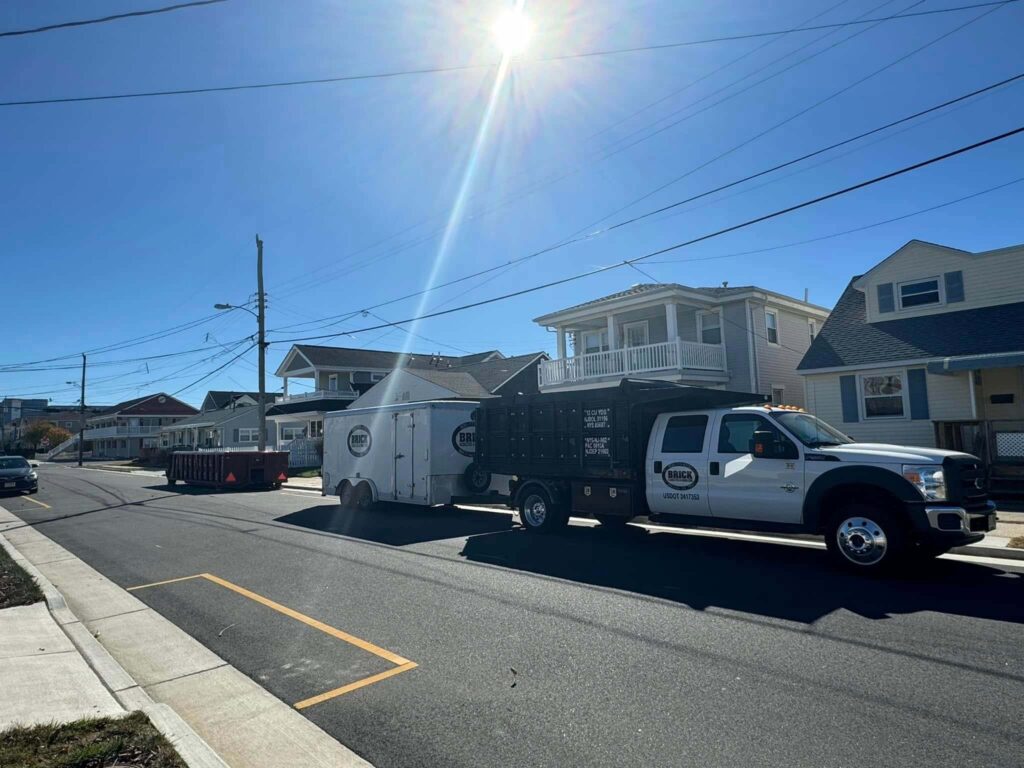 A work truck and trailer parked on a sunny residential street, with houses and utility poles in the background.