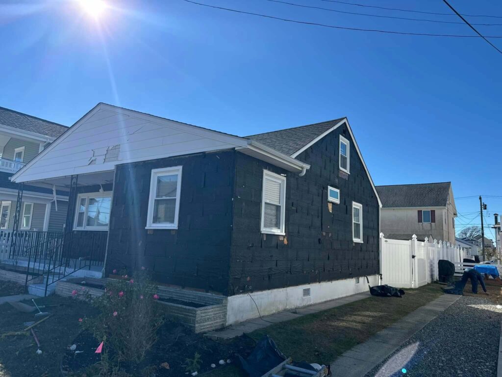 Side view of a black house undergoing renovations, with a sunny sky and neighboring homes visible in the background.
