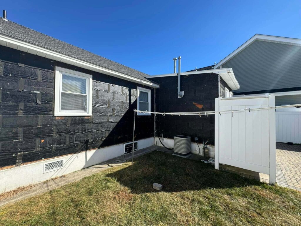 Side view of a black-painted house with white trim, featuring a grassy yard, a clothesline, and a patio area in clear blue sky.
