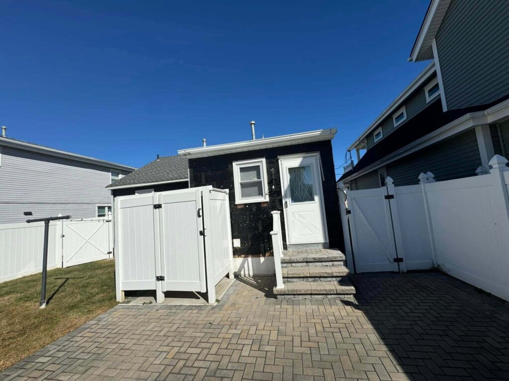 Small black building with white door and fenced yard, surrounded by other homes under a clear blue sky. Paved walkway leading up.