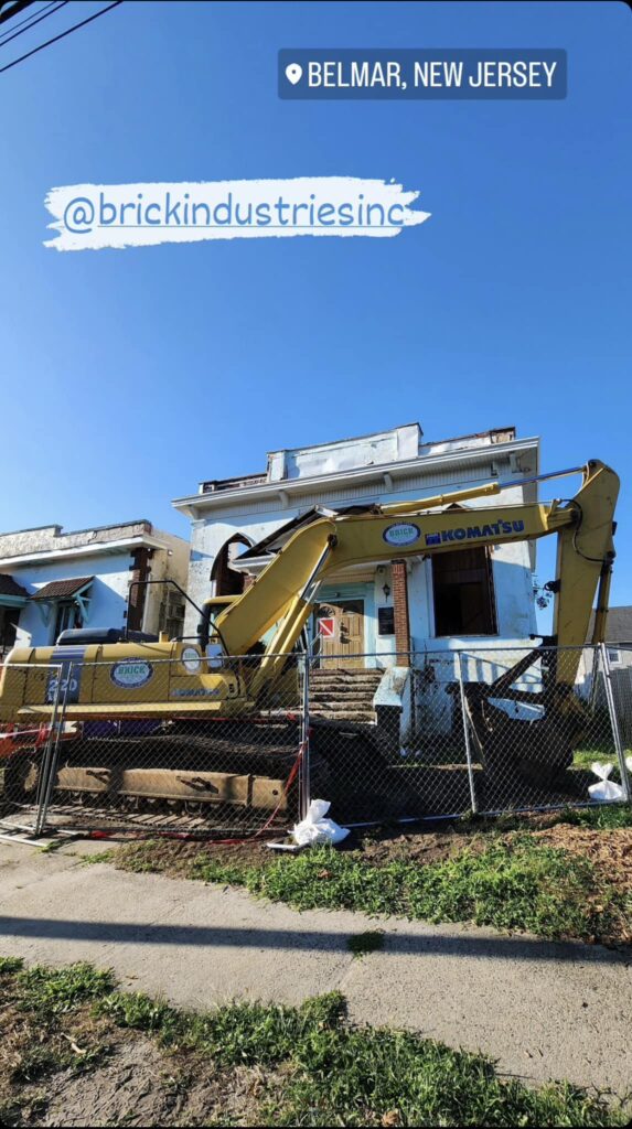 A yellow Komatsu excavator parked outside a partially demolished building in Belmar, New Jersey, with a construction site fence.