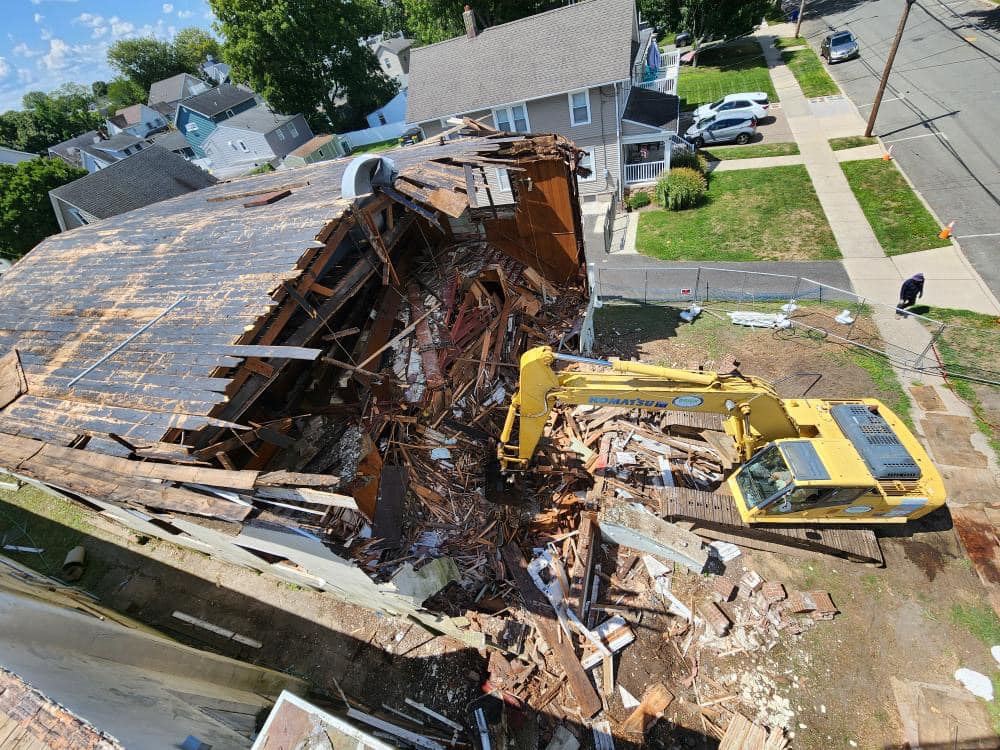 Aerial view of a damaged building being demolished by a yellow excavator, debris scattered on the ground in a residential area.