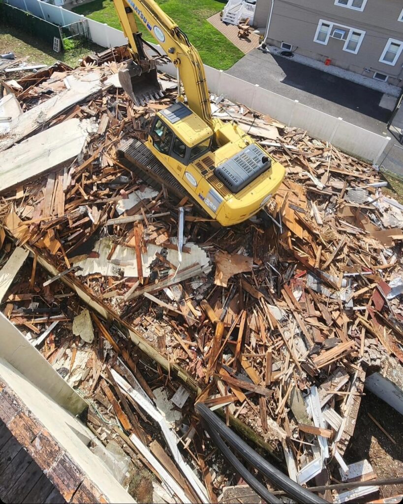 A yellow excavator on a pile of construction debris, surrounded by wood and building materials, near a house and green lawn.