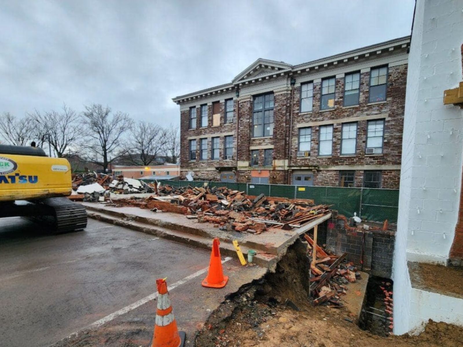 Construction site with heavy machinery near a partially collapsed building, surrounded by debris and safety cones on a cloudy day.