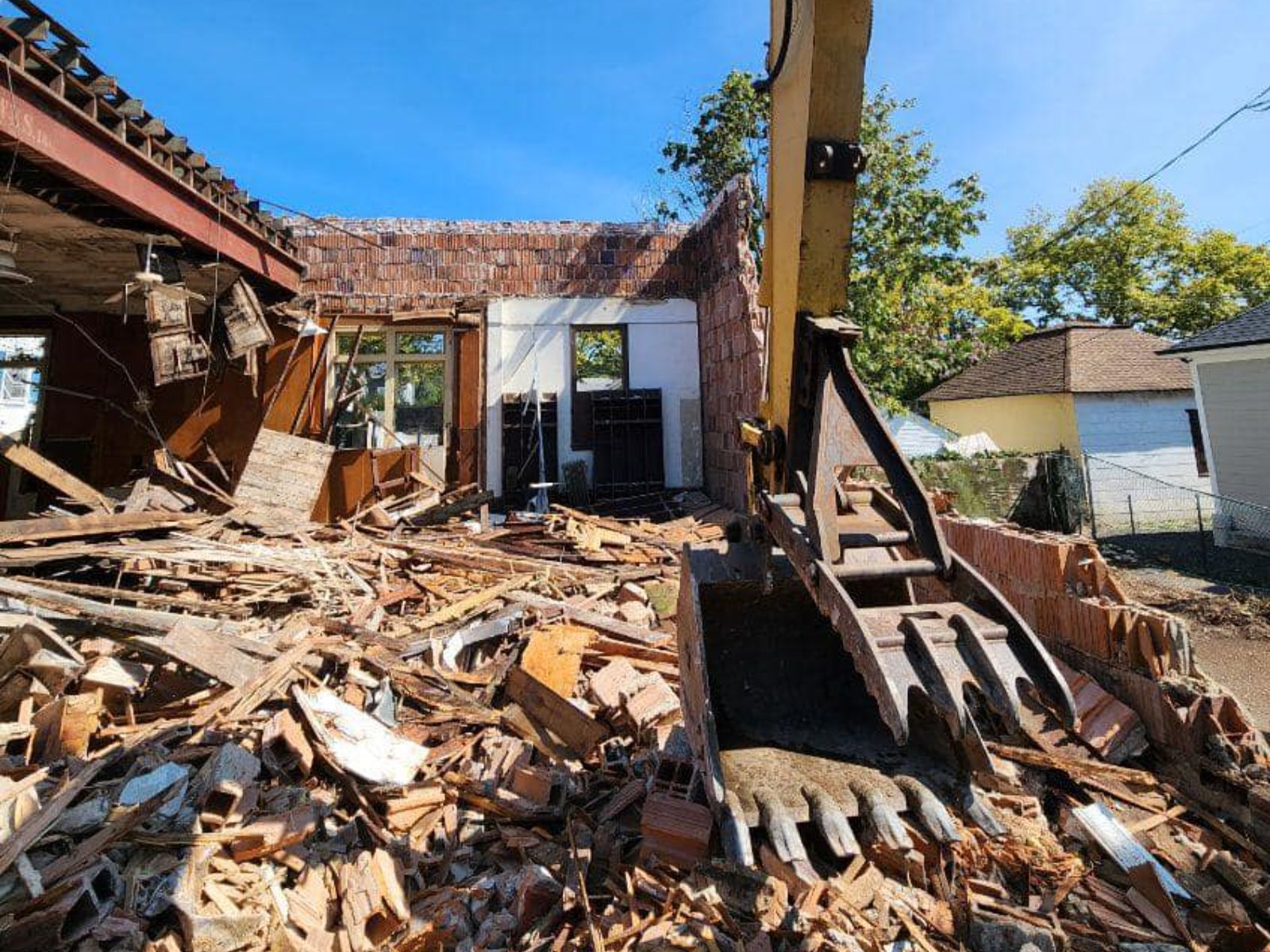 A demolition site shows debris scattered around, with a construction machine actively breaking down a structure under a blue sky.