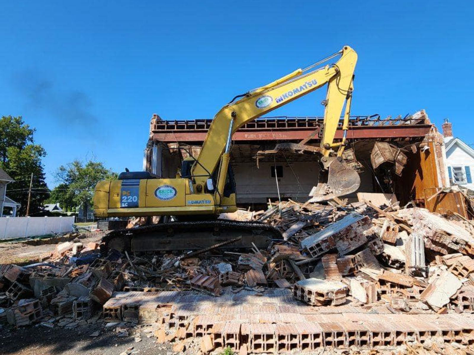 Excavator demolishes a structure, creating a large pile of debris amid a residential area with trees and houses in the background.