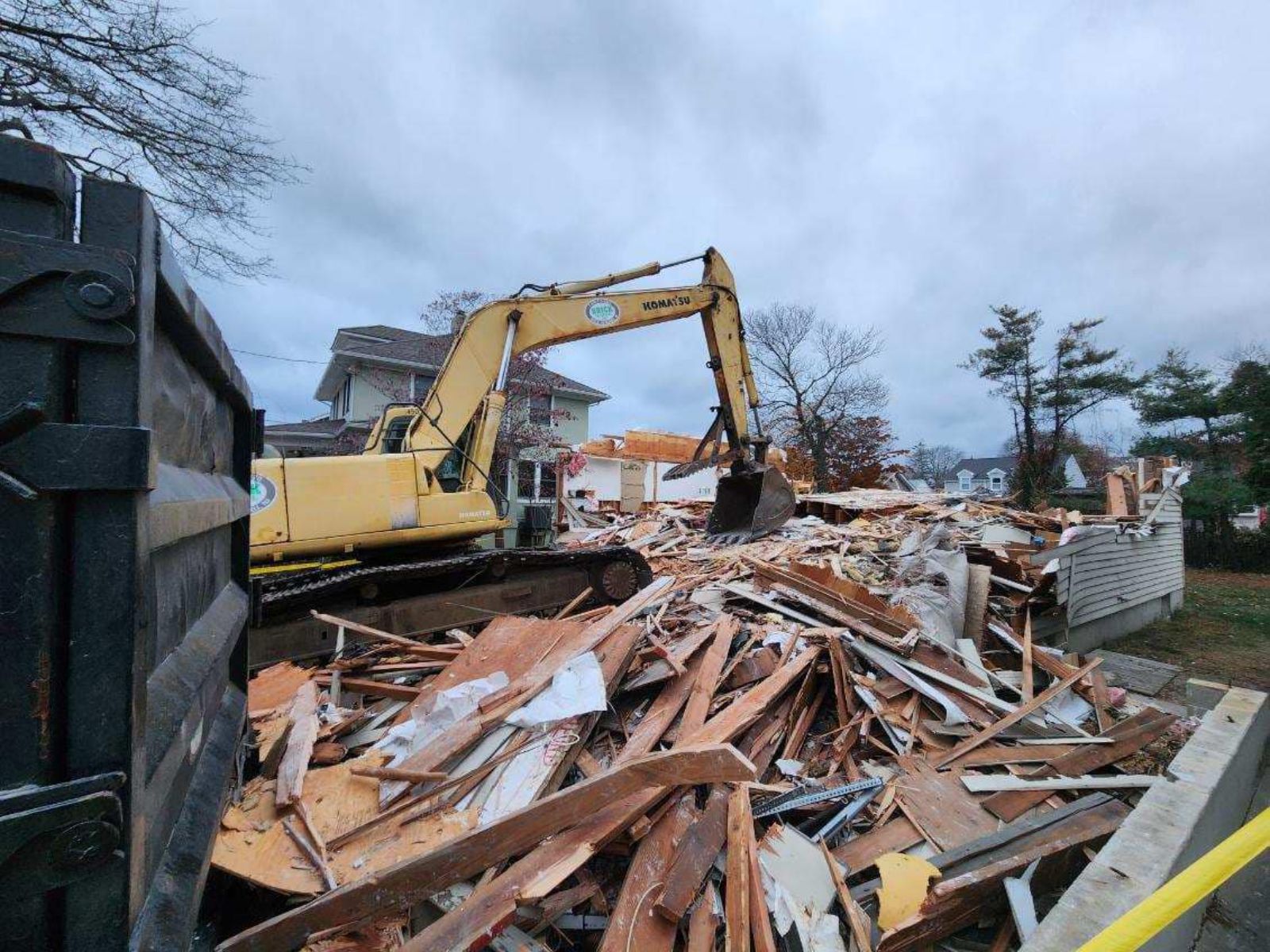 A yellow excavator demolishes a structure, surrounded by debris, under a cloudy sky in a residential area.