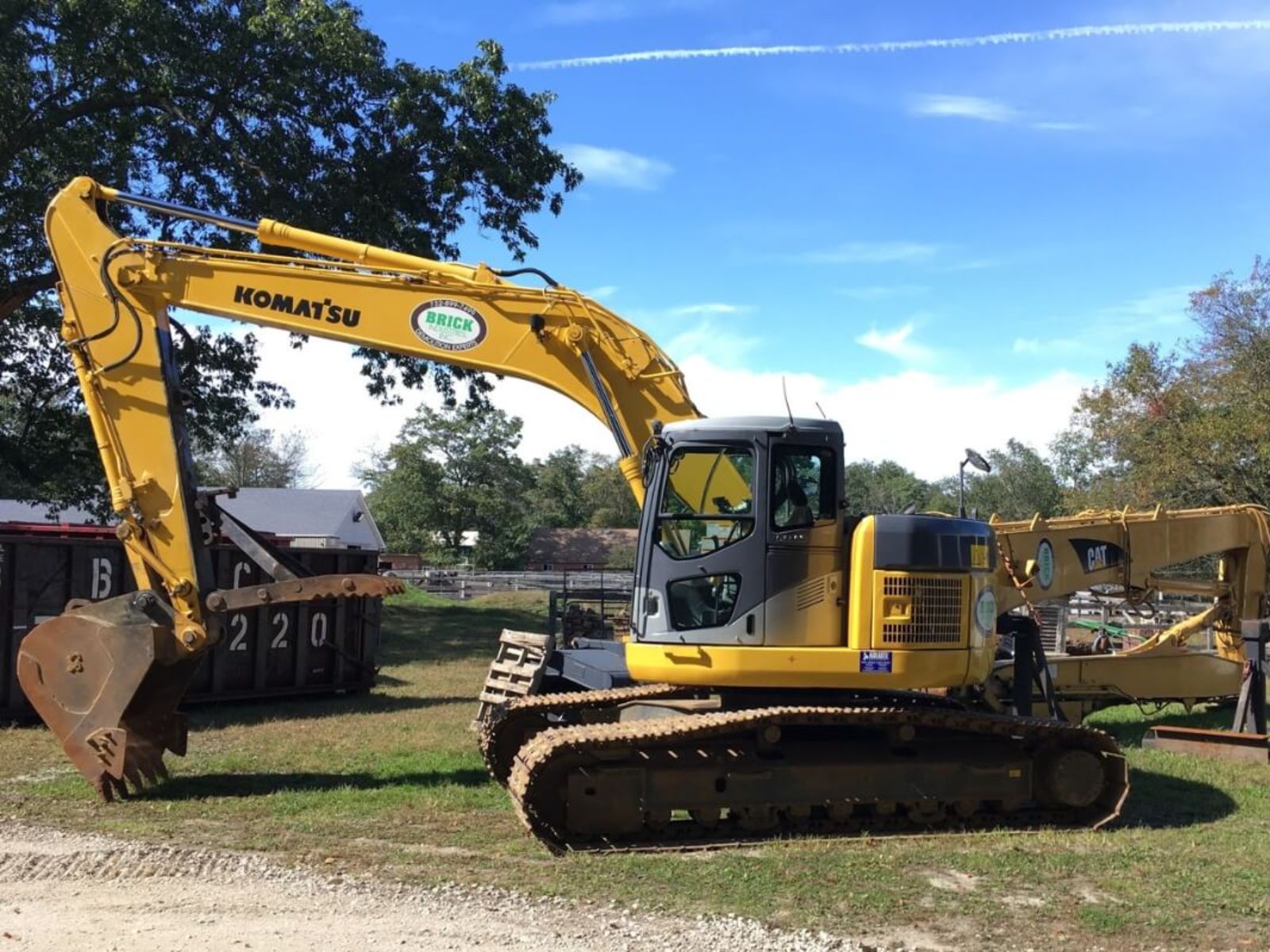 user-inserted A yellow Komatsu excavator with a large digging bucket parked outdoors against a blue sky and green surroundings.