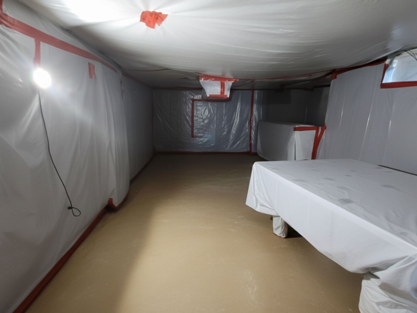  A room featuring a table covered with plastic and a white sheet, suggesting preparation for an event or cleaning.