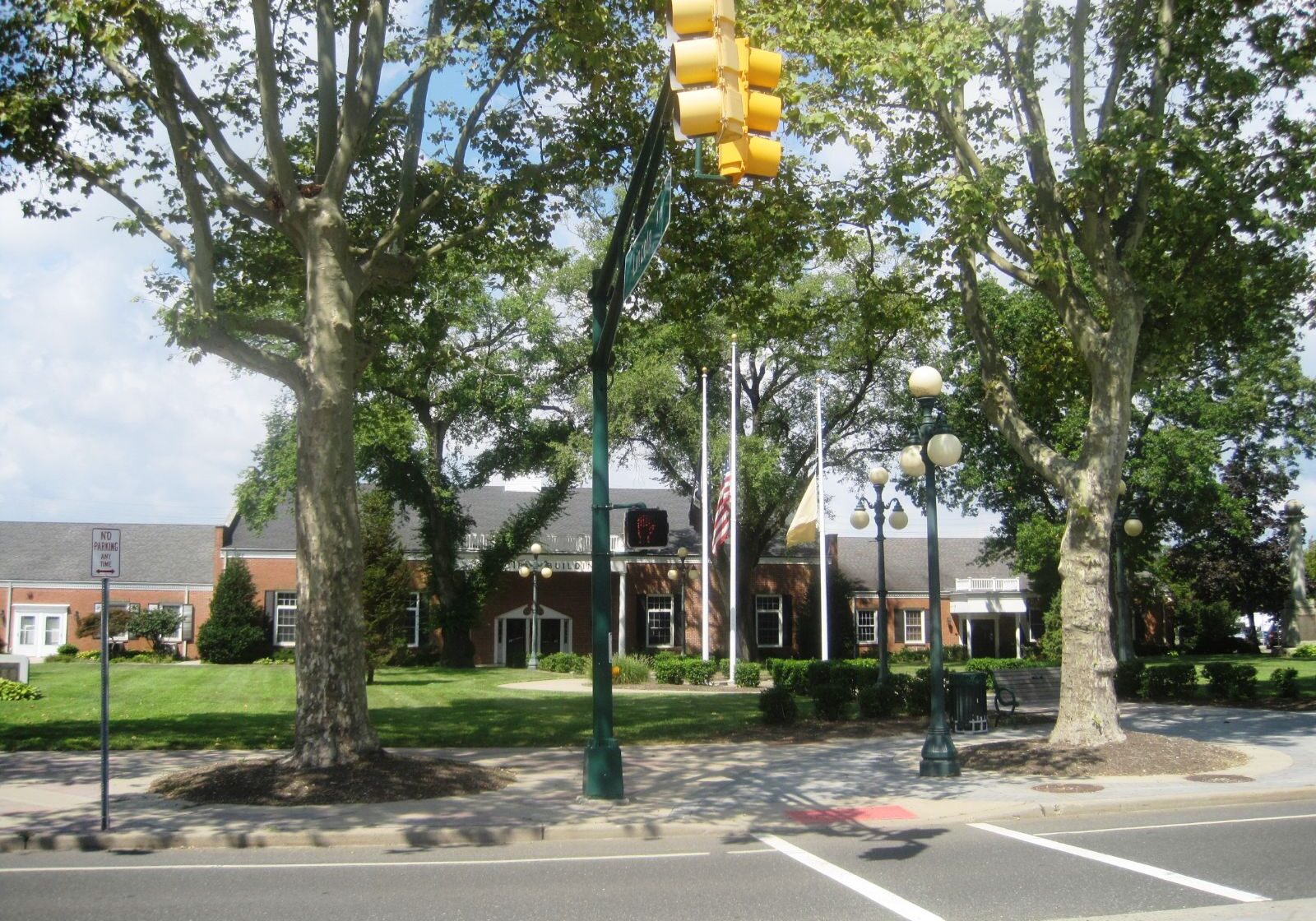 Avon-by-the-Sea downtown street view with large trees, a traffic light, and a well-maintained community building with flags in the background.