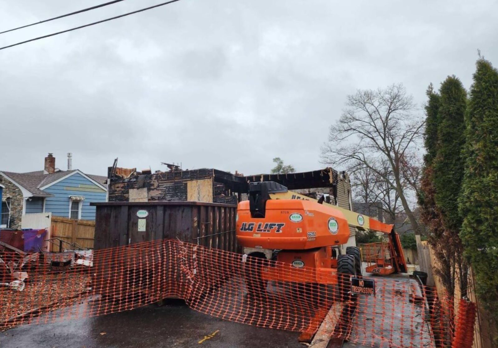 Construction site featuring a damaged building with a lift, debris, and orange safety fencing under a gray sky.