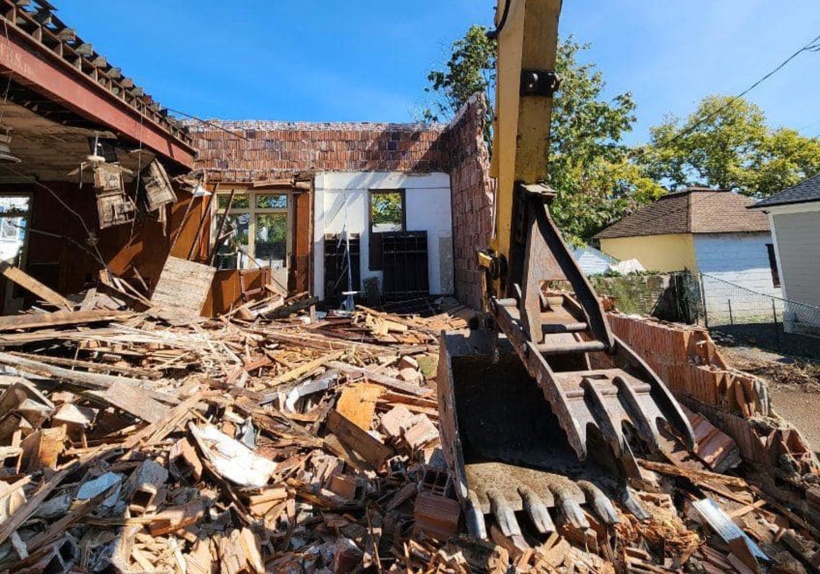 A demolition site shows debris scattered around, with a construction machine actively breaking down a structure under a blue sky.