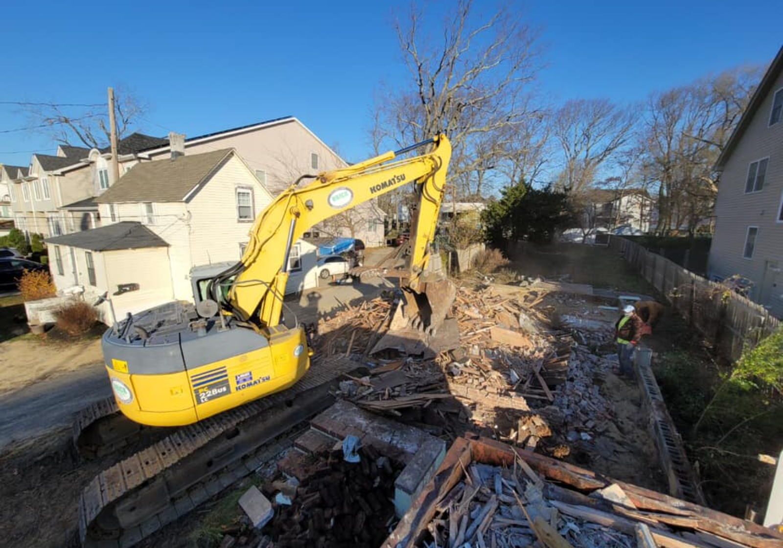 Excavator demolishing debris in a residential area, with workers nearby and clear blue sky in the background.